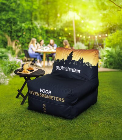 Een zwarte Old Amsterdam-zitzak met goudkleurige grachtenpand-skyline en de tekst 'Voor levensgenieters', in een zomerse tuin naast een bijzettafel met kaas.