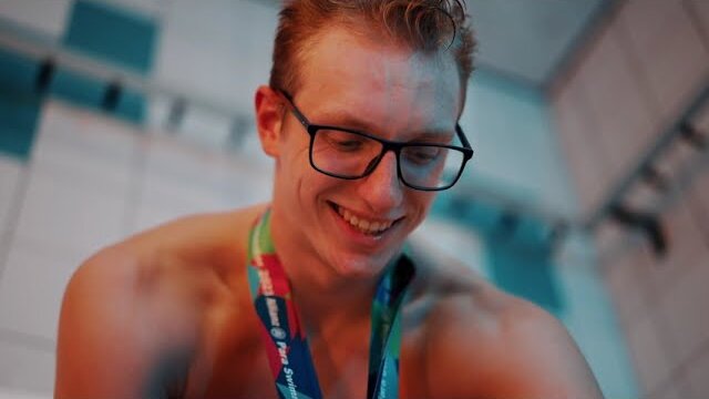 Stilstaand beeld uit de bedrijfsvideo van BeGlobal: een lachende zwemmer met bril en een medaille om de nek, aan de rand van het zwembad.