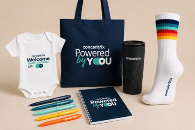Concentrix-merchandise op een beige achtergrond: een rompertje met 'Welcome little game-changer', een donkerblauwe tas en notitieboek met 'Powered by You', een zwarte thermosbeker, een witte sportsok met regenboogstrepen en zes gekleurde pennen.