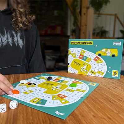 Het bordspel Dierenredders opengeslagen op een houten tafel, met de doos rechtop erachter en een speler die een pion verzet.