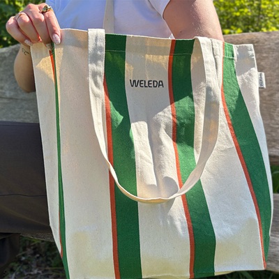 Een groen-oranje gestreepte katoenen shopper met WELEDA-opdruk, in de hand gehouden in het zonlicht.