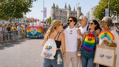 Vijf lachende bezoekers van Antwerp Pride op straat voor Het Steen, met merchandise uit de collectie: een wit en een regenboogkleurig rugzakje met AP-logo, een jute draagtas, een zwarte drinkfles, polsbandjes, een regenboogwaaier en een knuffel aan een riem. Op de achtergrond een praalwagen met regenboogvlaggen en publiek achter hekken.