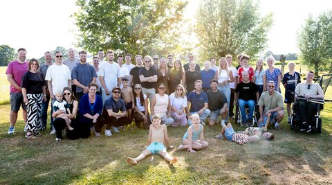 Het team van PinkCube met partners en kinderen, samen op een grasveld voor een rij bomen tijdens de zomerse bedrijfs-BBQ.