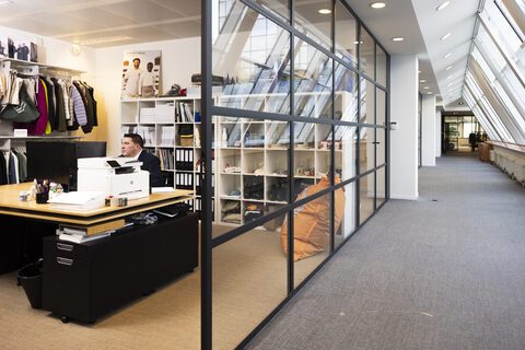 Het kantoor van Wazabi: Julien Grandjean aan zijn bureau, met achter de glazen wand de showroom met stalenrekken en een rek met textiel.