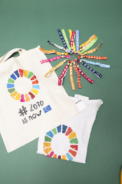 Een linnen tote bag met het SDG-kleurenwiel en de tekst '#2030 is now' naast een grijs T-shirt met hetzelfde wiel en een waaier van gekleurde polsbandjes.
