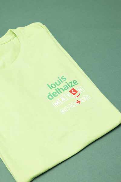 Een opgevouwen lichtgroen T-shirt met op de borst de logo's van Louis Delhaize, Match en Smatch.