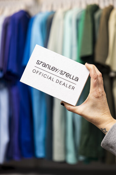 Een hand houdt de kaart 'Stanley/Stella Official Dealer' omhoog voor een rek met textiel in blauwe en groene tinten.